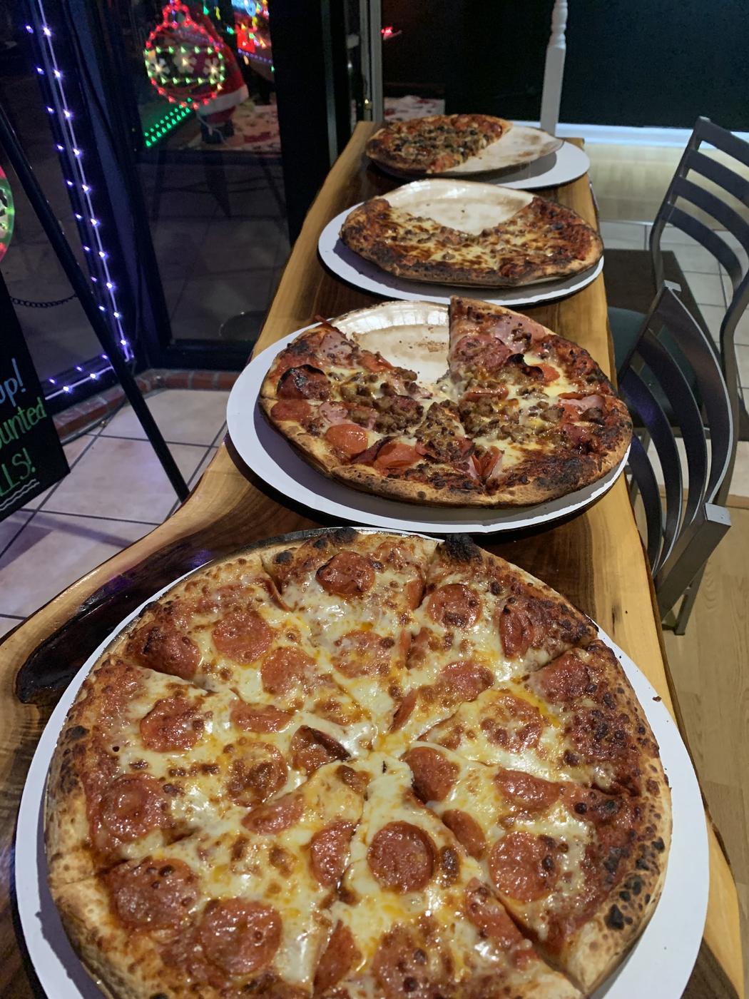 Four pizzas on white plates lined up along a live edge wooden bar top, with lights in the window behind
