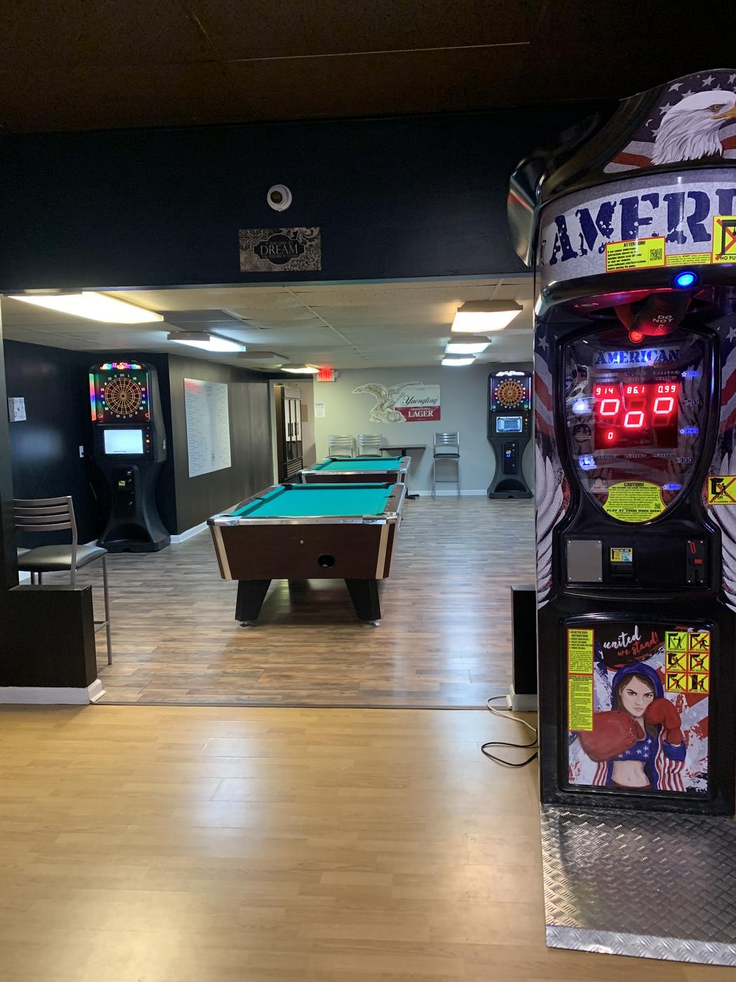 The back room with two pool tables in teal cloth, two lit dart machines, a boxing arcade machine in the foreground and a tournament bracket board on the wall